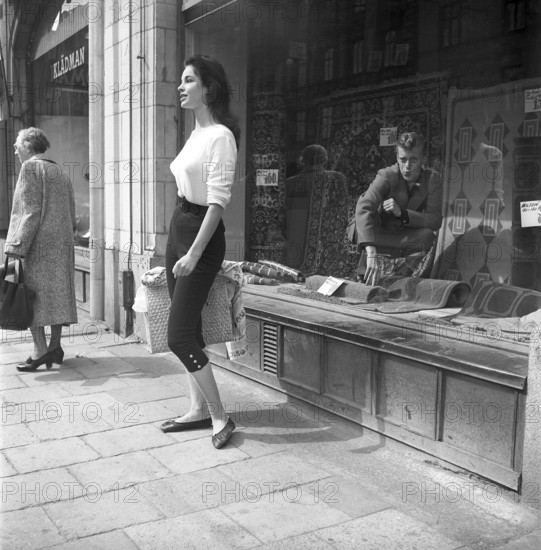 Young woman in 1956. She is standing in front of a shop window on Strandvägen in Stockholm and maybe she is not aware of the guy in the carpet shop who is sitting inside the window and looking at her admiringly. She is wearing a white jumper and knee-high trousers. Sweden 1956. Kristoffersson ref BS94-3