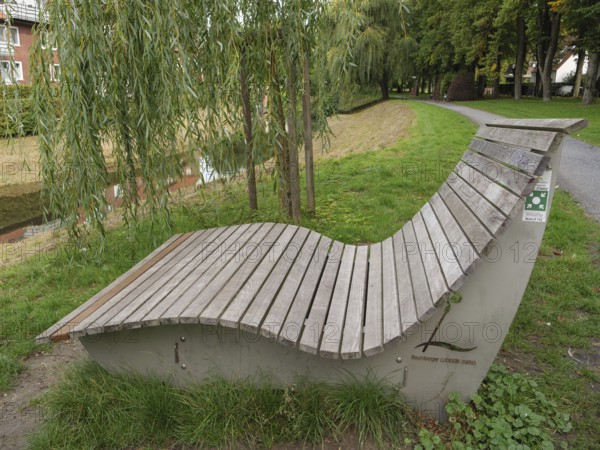 A curved wooden bench stands under a willow tree along a park path, suitable for relaxation, Coesfeld, münsterland, germany