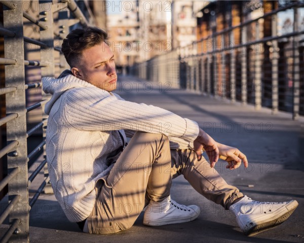 Handsome trendy young man, sitting in city setting at sunset wearing a fashionable sweater or cardigan