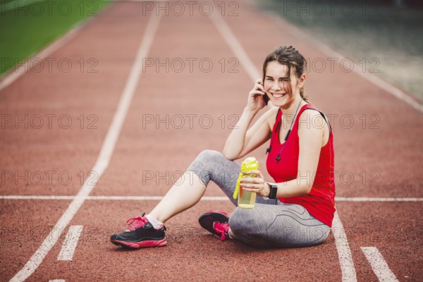 Sport and technology. A beautiful young Caucasian woman with ponytail sitting resting after workout during run at stadium, a red treadmill track. Uses for cell phone headphones and sports watches