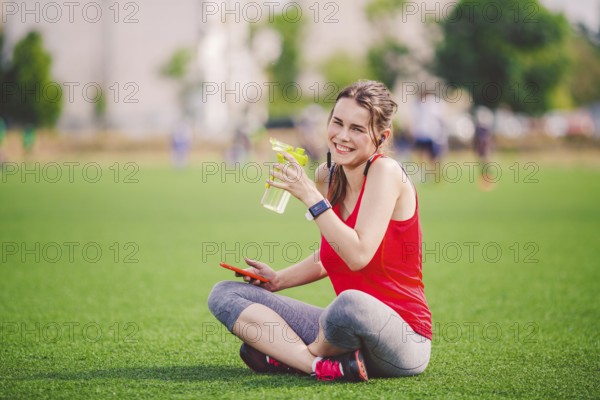 Theme sport health. beautiful young girl sitting resting on green grass lawn stadium using technodogies. In handphone in ear headphones listens music, summer sportswear smart watch heart rate monitor