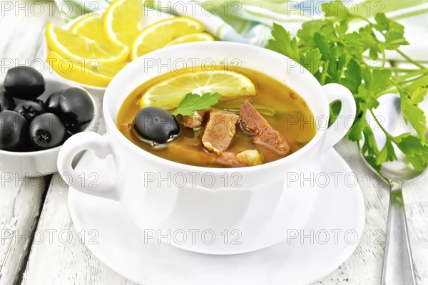 Soup saltwort with lemon, meat, pickles, tomato sauce and olives in a white bowl, towel, parsley on a light wooden board background