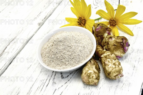 Flour of Jerusalem artichoke in a white bowl with yellow flowers and vegetables on the background of wooden boards
