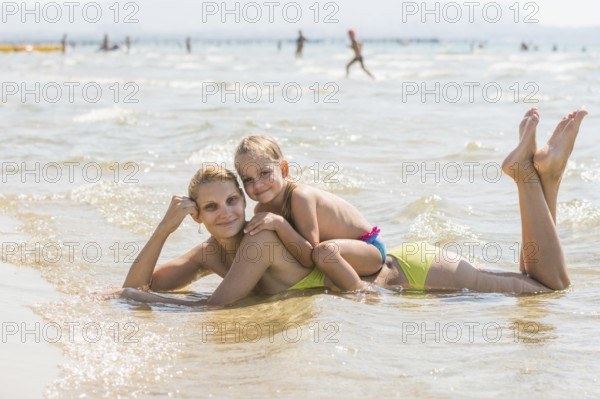 Mother and her sitting on the back of a four-year girl lying in the water on the sandy beach and happily look into the frame
