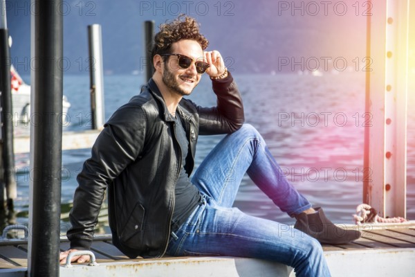 Handsome man on a lake in a sunny, peaceful day, sitting on a wood pier, thinking or meditating
