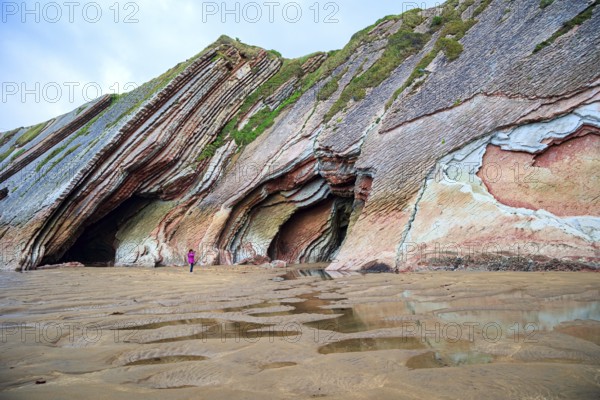 Picturesque cave on the beach. Flysch - underwater rocks exposed at low tide. Low tide in the Atlantic. Woman in pink jacket photographs Playa Zumaya