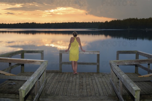 Young woman standing on a pier and admired the sunset on a lake in Småland, Sweden, Europe