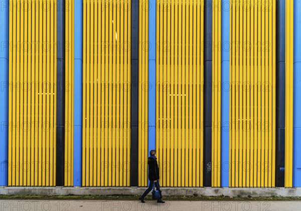 Pedestrian woman walking in front of a colorful Industrial background with straight patterned vertical lines, Brussels, Belgium, Europe