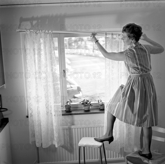 1960s home decorating. A woman at home putting up the curtains in the room window. All by herself she balances on a chair and a stool to manage it. Sweden 1966