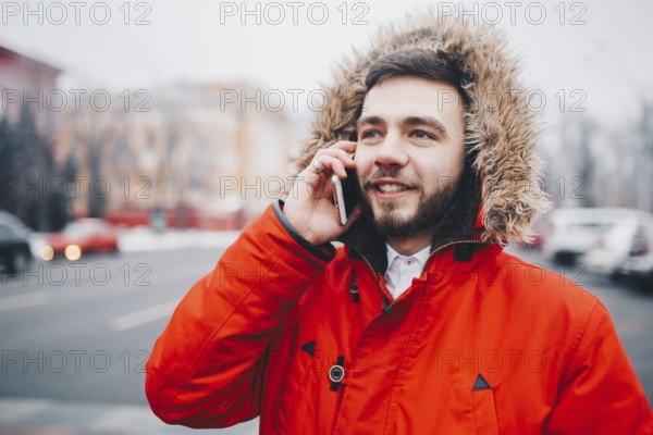 Young guy with beard and red jacket in hood a student uses mobile phone, holds in his hand near the head, talking on the phone with smile on the background of red building the university or college