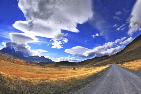 Gray dirt road in the Chile National Park Torres del Paine. Incredible shaped cloud formed by glaciers glisten in the sun. On the horizon are seen mountains with snow-capped peaks. Picture taken with a fisheye lens