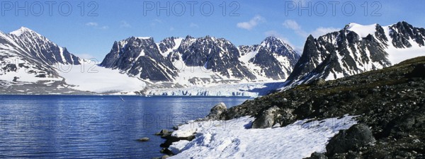 Landscape on the Magdalenenfjord, which is bordered by snow-covered, rugged mountains. At the end of the fjord, glaciers push their ice directly into the dark blue water of the fjord. Bright sunshine, blue sky, deserted. Panorama format. Landscape at Magdalene Fjord, bounded by snow-covered, rugged mountains. At the end of the fjord glaciers push their ice directly into the deep blue waters of the fjord. Panorama view