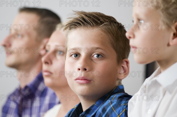 Close-up of a seven-year-old male teenager with parents lined up in the blurred background and brother looking seriously into the camera