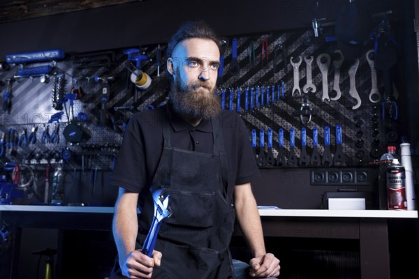 Portrait of small business owner of young man with beard. Guy bicycle mechanic workshop worker sitting with tool in his hand in a working black clothes in an apron in the background of a bicycle shop