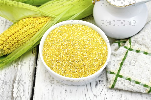 Corn grits in a white bowl, cobs, a jug of milk and a napkin on a wooden boards background
