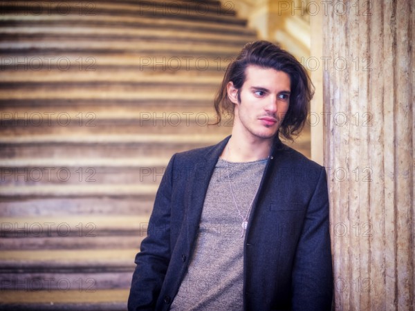A young, long-haired attractive man standing next to a marble column, leaning onto it, wearing elegant jacket