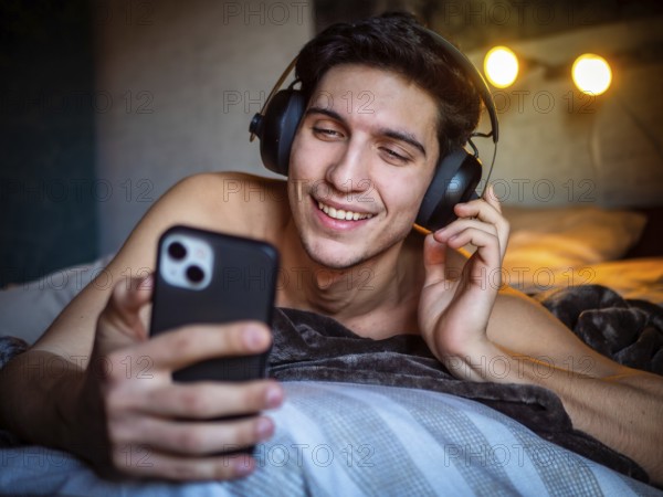 Glad male in headset smiling and text messaging on mobile while listening to favorite song and relaxing on bed on weekend