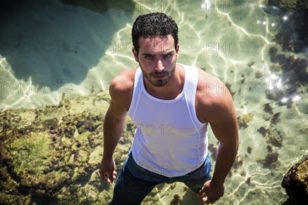 Attractive young athletic man in the sea or ocean by the rocky shore, wearing wet t-shirt, serious expression