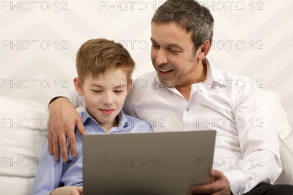 Father and son sitting on the sofa at home. Happy dad embracing boy and watching him working with laptop