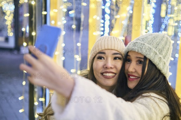 Two girlfriends asian girls in white coats are having fun hugging and selfie on mobile phone, new year, light bulbs