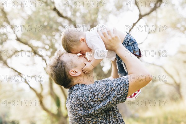 The father holds his son above him, they touching their noses, the son holds the father by the cheek .