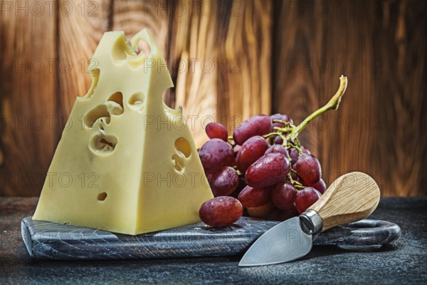 Piece of maasdam cheese on vintage carving board knife and bunch of rose grape old wooden background