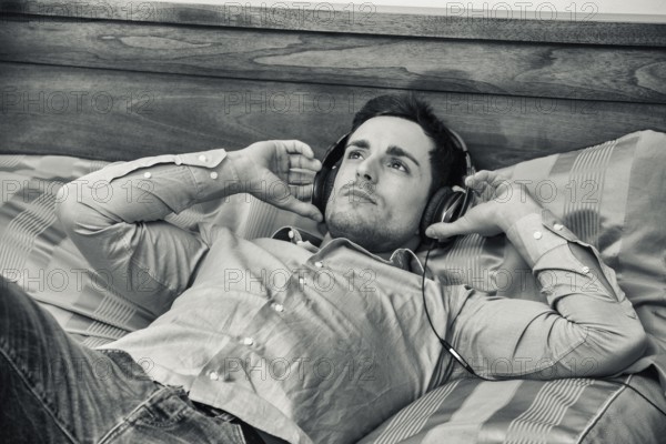 Handsome young man in bed listening to music with headphones, using cell phone or MP3 player