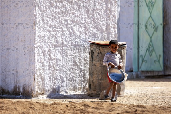 A child stands with a bucket in the sun next to a white house wall, Little boy stands by the house with a bowl in his hand in the Sahara in Algeria