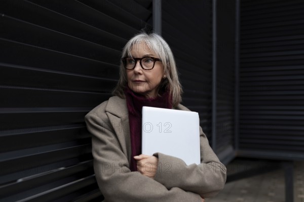 A middle-aged businesswoman with glasses is standing against a dark wall, holding a laptop. She looks thoughtful and confident, dressed in a stylish coat
