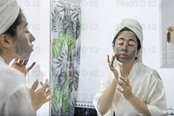 Portrait of a young woman applying a black cream face mask for skin in front of her bathroom mirror after a relaxing shower. Skin care and female beauty. Daily routine