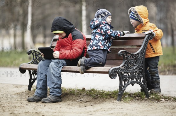 Three young boys playing on a park bench in winter wrapped up warmly against the cold weather with one youngster reading a tablet as the two younger children romp around