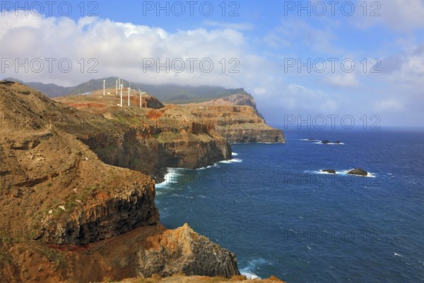 Eastern tip of the island of Madeira. Rocks steeply in the blue waters of the Atlantic Ocean. Over a cliff on the ocean breeze are the windmills