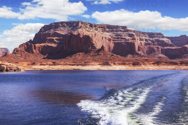 Foamy trace of motorboat crosses the emerald waters. Red sandstone hills surround the lake. Lake Powell on the Colorado River