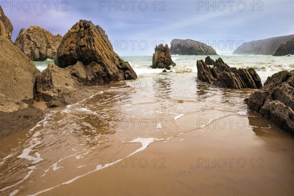 The smooth wet sand of the beach reflects the rocky boulders. The Playa de la Arnia beach. Snow-white sea foam glistens. Spain, Cantabria