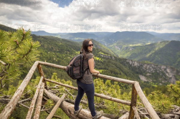 Young tourist woman with backpack enjoys the view from a wooden bridge high in the mountain designed for viewing the side from above. In pine trees. Vivid image of the mountain. Tourism destination