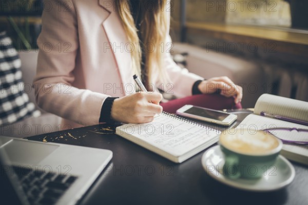 Close up caucasian woman hand on wooden table inside cafe makes notes in notebook. Subject freelancer blogger journalist at work. Unrecognisable person. Technology phone and laptop with cup coffee