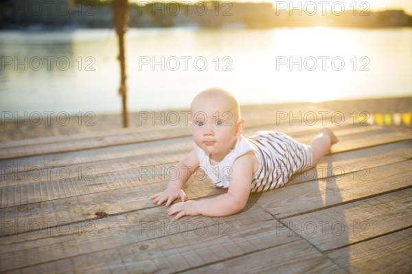 A child a boy, one year old, blond man lies on his stomach on wooden dock, pier in striped clothes, compound near pond on sandy beach against background of river in the summer at sunset of the day