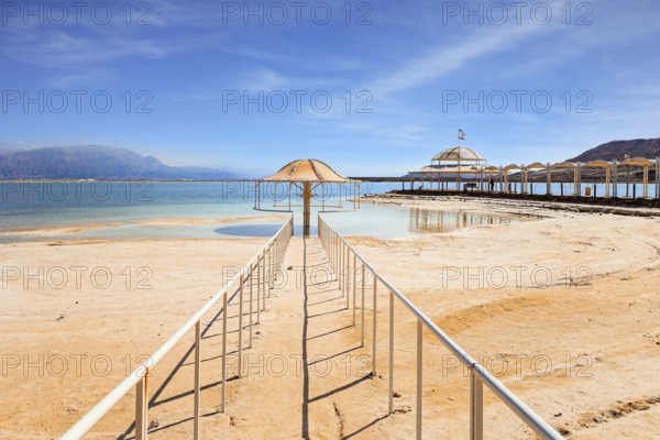 Railings enclose the path to the water. Lovely round umbrella protects from the sun. The drainless salt lake in the Middle East. Dead Sea. Coast of Israel
