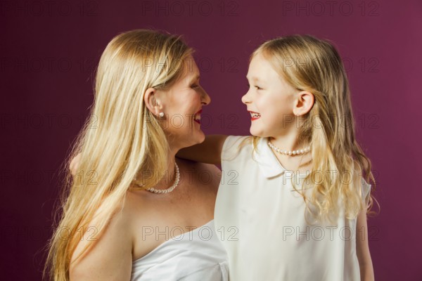 Portrait of happy smiling mother and daughter, both with long loose blond hair, wearing white clothes and red lipstick. Daughter looks like mother. Studio with purple background