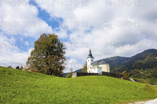 White church and bell tower on a hill near the small village. Green grassy lawns in a mountain valley. Travel to Slovenia. Picturesque Julian Alps