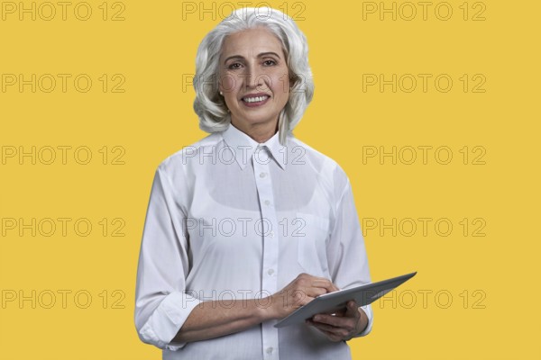 Smiling mature business woman holding pc tablet and looking at camera. Beautiful elderly woman with digital tablet pc on yellow background