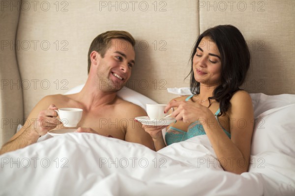 Couple holding cups in bed. Young man smiling. You keep surprising me