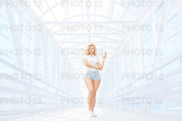Beautiful young woman with music headphones standing on the bridge with a takeaway coffee mug and posing in front of an urban background