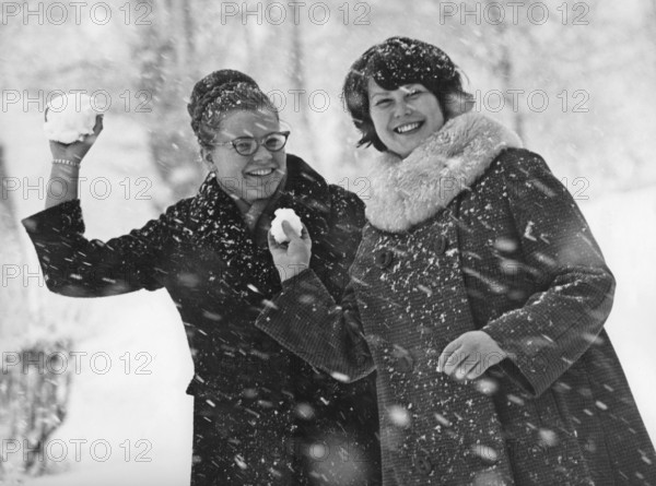 Winter in the 1960s. Two young women is aiming and about to throw snowballs at the photographer. The snow is falling and they are dressed warm with winter coats. Sweden 1964