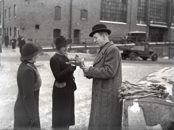 In the 1940s. Times are tough and the unemployed man is trying to make a living with temporary jobs. Pictured here in a street in Stockholm when selling tulips. Two women has stopped to buy some from him. Sweden 1940 Kristoffersson ref 37-9