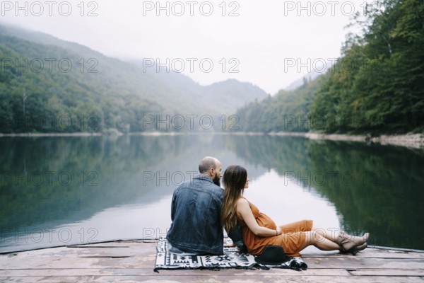 Man with pregnant woman are sitting on a pier by the lake in the park.