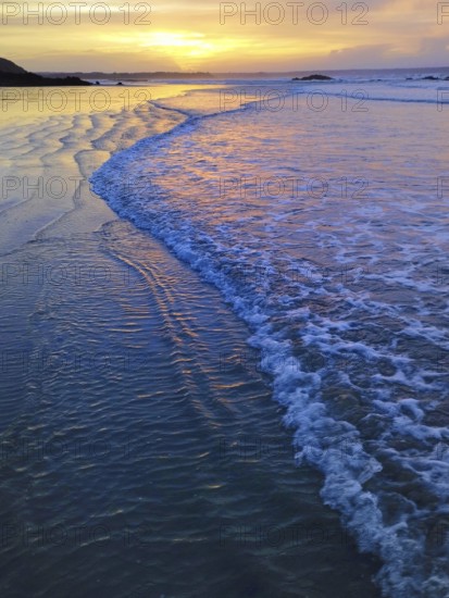 A serene beach scene with gentle waves reflecting a vivid sunset sky, creating a calm and peaceful atmosphere. Planguenoual