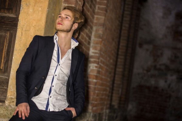 Handsome blond young man standing against brick wall, looking up to a side, wearing white shirt and jacket