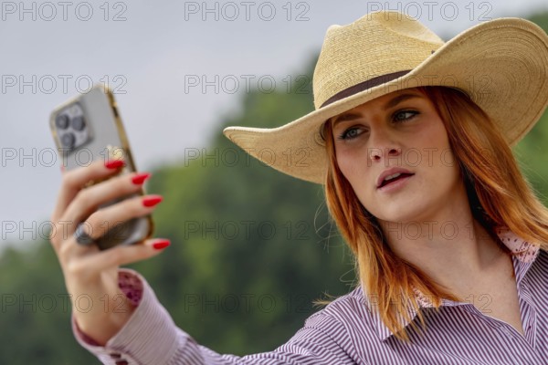 A beautiful redhead model uses her cellphone while posing in a country setting wearing cowgirl attire
