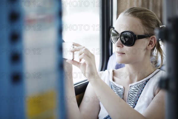 Attractive young woman wearing sunglasses holding up and photographing at her mobile on a bus or train